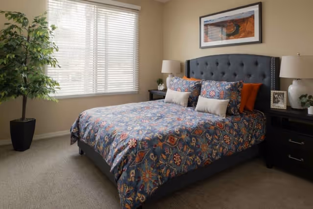 Sunlit bedroom with a patterned bed and tufted headboard, nightstands with lamps, a framed artwork, and a potted plant by the window.