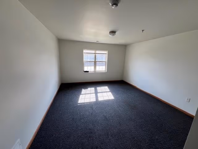 Empty room with white walls, a single window letting in natural light, and dark carpeted floor. The room has a simple ceiling with two light fixtures and wooden baseboards along the walls.