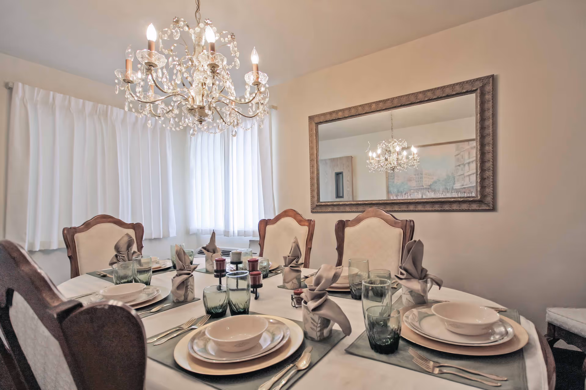 A formal dining room with a round table set for six people. The table is covered with a white tablecloth and has place settings including plates, bowls, silverware, green-tinted glasses, and folded napkins. Above the table hangs an ornate crystal chandelier. A large framed mirror is mounted on the wall, reflecting the chandelier and part of the room. White curtains cover the windows, allowing soft natural light to fill the room.