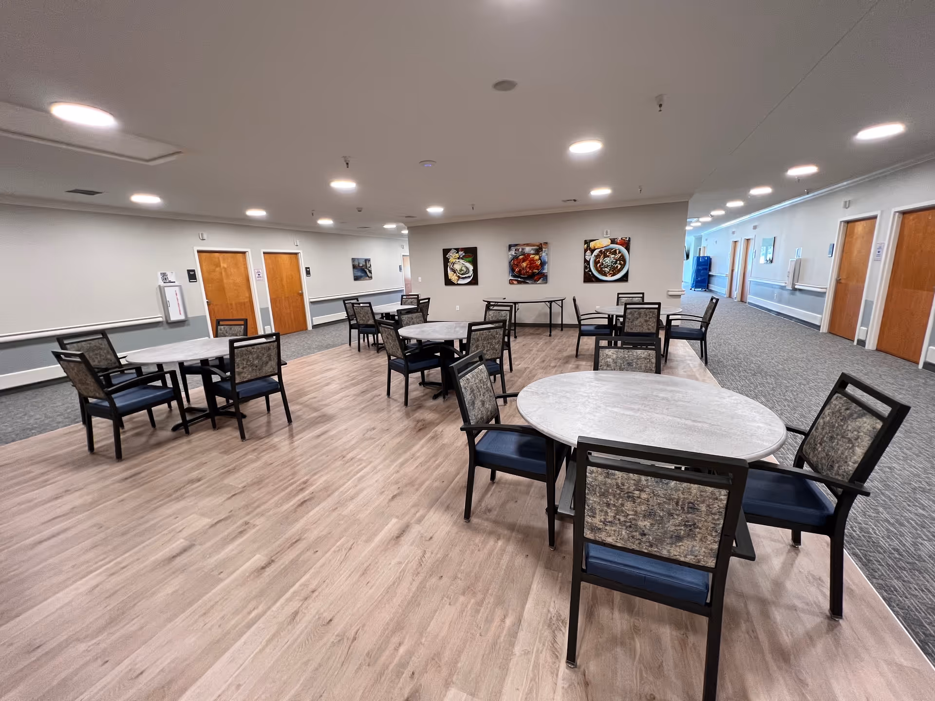 Spacious communal dining area with round tables and chairs and a long hallway in a senior living facility.