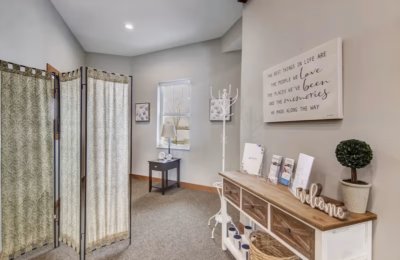 A cozy interior corner of a senior living facility with a decorative folding screen on the left, a small table with a lamp and decorative items under a window, a white coat rack, and a wooden console table with drawers and brochures. A potted plant and a wooden 'welcome' sign are on the console table, and a wall art piece with an inspirational quote hangs above it.