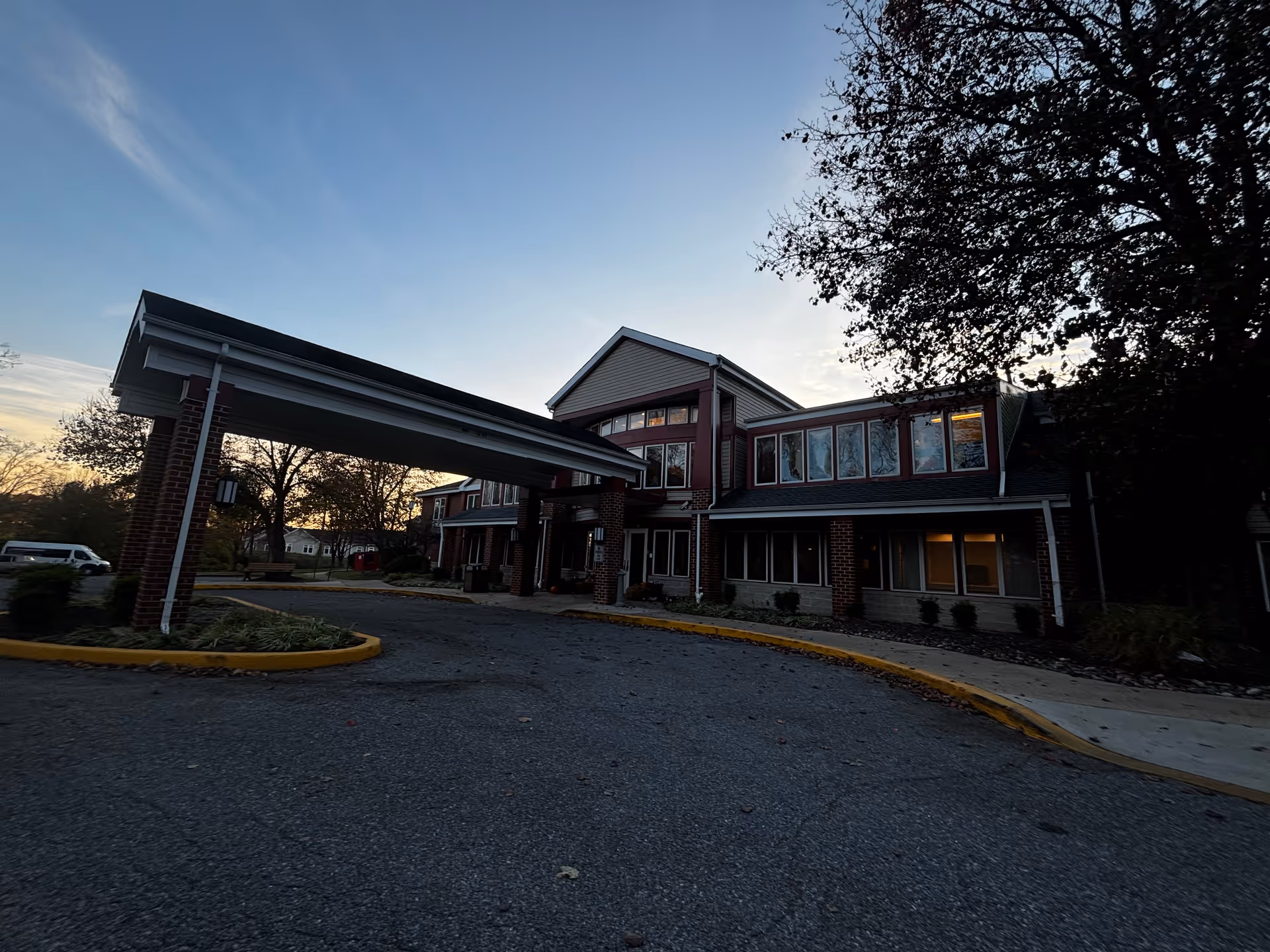Exterior view of a two-story senior living facility building with a covered entrance driveway, surrounded by trees and a parking area at dusk.
