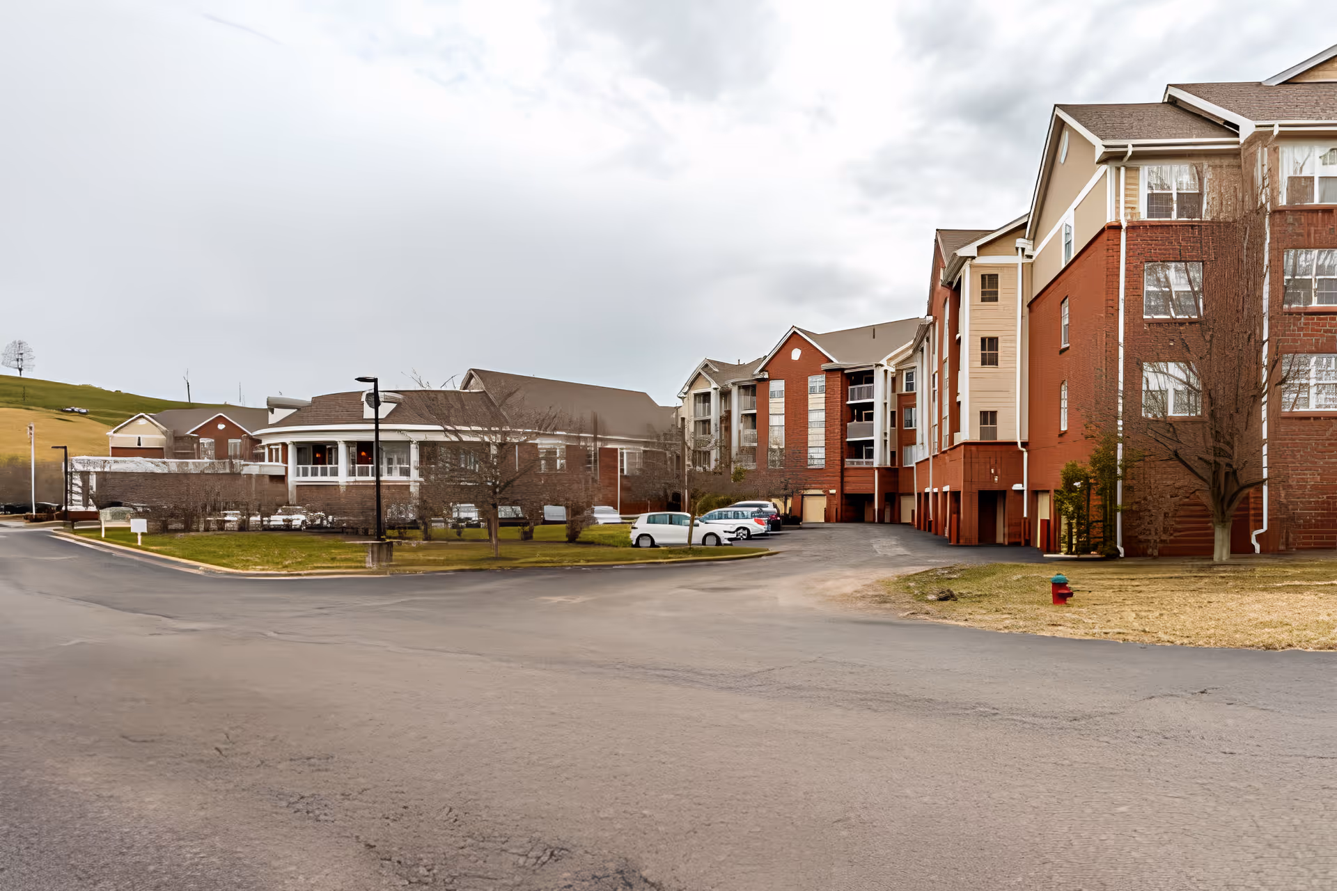 Exterior view of a multi-story red brick and beige senior living facility with a parking area, lawn and cloudy sky.