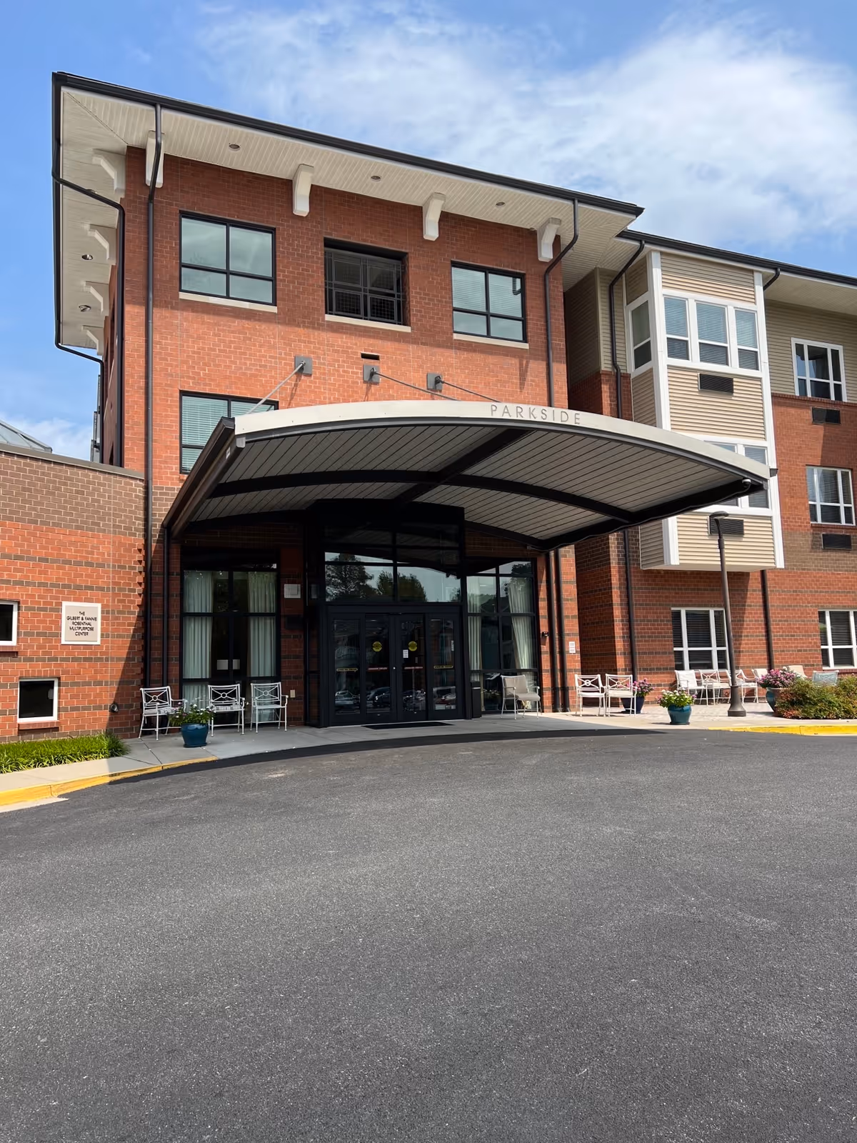 Front entrance of a three-story brick senior living facility with a large curved canopy labeled 'PARKSIDE' and chairs on the entry patio.