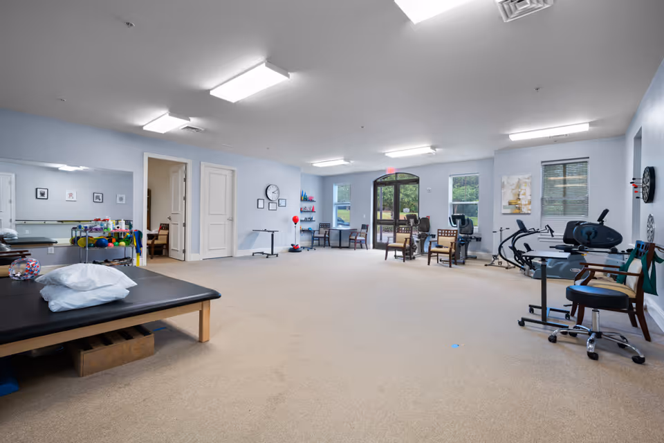 A spacious physical therapy or rehabilitation room with exercise equipment, chairs, therapy tables, and various therapy tools. The room has light blue walls, large windows, and bright overhead lighting. There is a door leading outside and a large mirror on one wall.