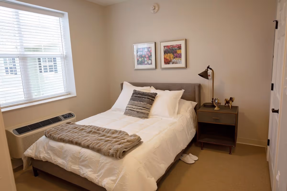 A neatly made bed with white bedding and a gray decorative pillow in a small bedroom. A folded beige blanket lies at the foot of the bed. Next to the bed is a wooden nightstand with a vintage-style clock, a small horse figurine, and a brass desk lamp. Two framed pictures of colorful flowers hang on the beige wall above the bed. A window with white blinds is on the left side, and a pair of white slippers is partially visible on the floor near the nightstand.