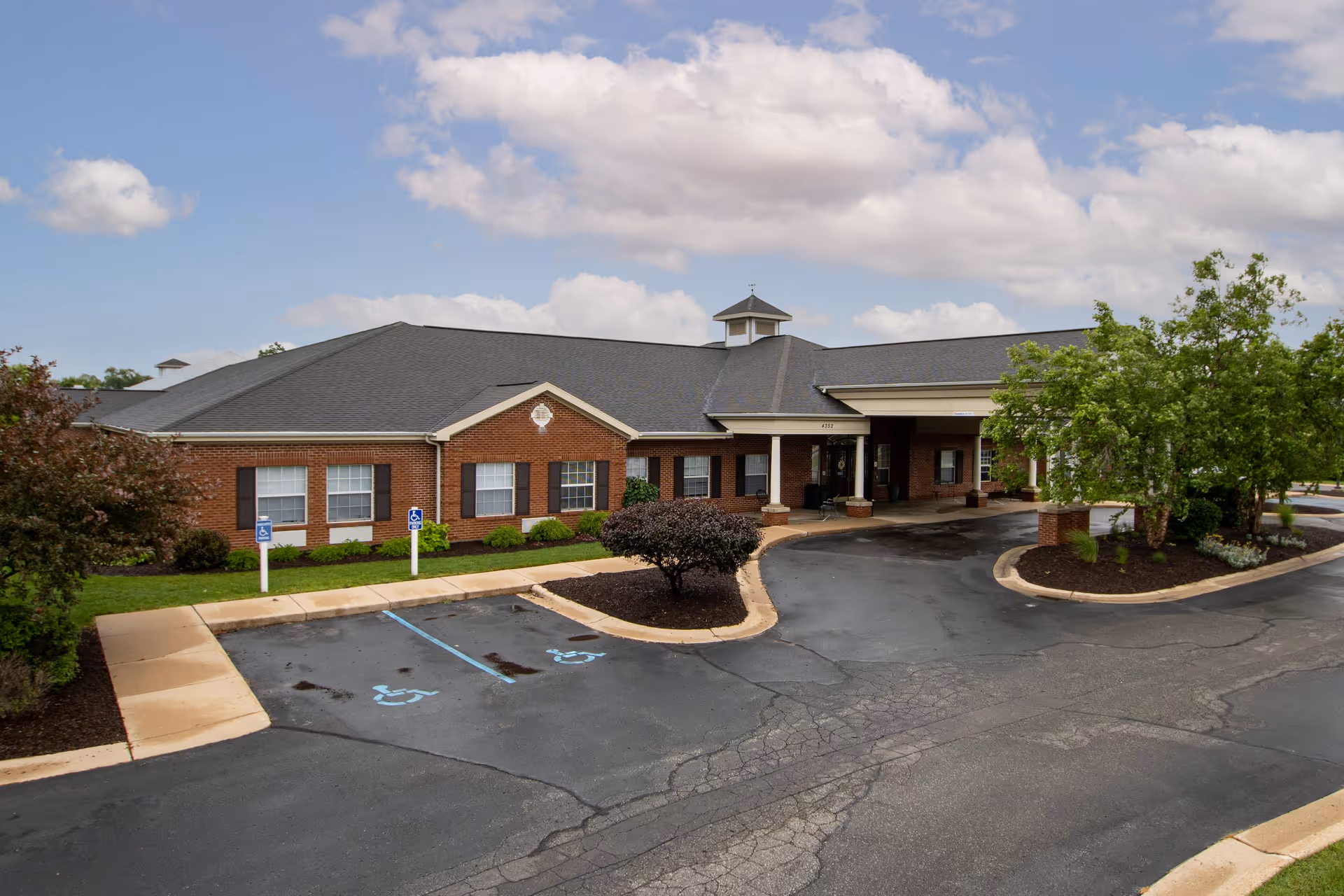 Exterior view of a single-story brick building with a dark shingled roof, surrounded by landscaped greenery and trees. There is a covered entrance with a driveway and two handicap parking spaces in front. The sky is partly cloudy.