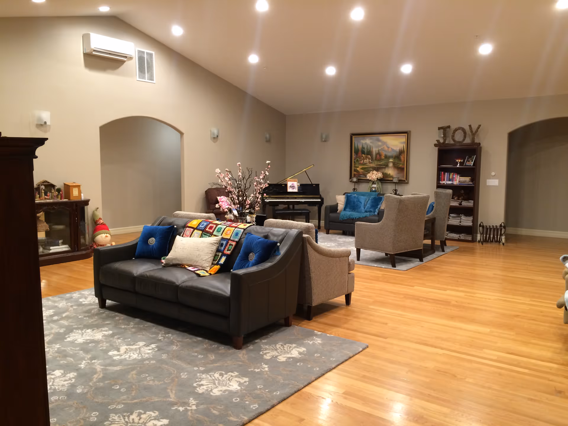 Spacious common living room with sofas, armchairs, a piano, bookshelf and hardwood floors.