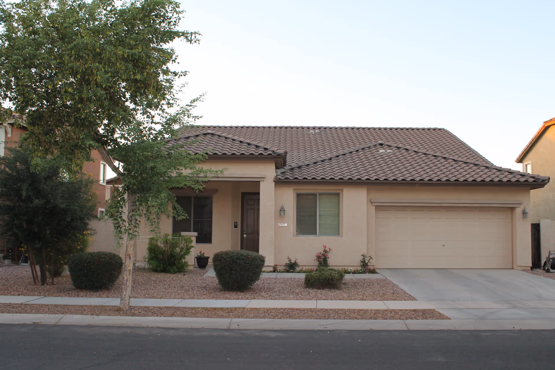 Single-story house with a tiled roof, beige exterior walls, a two-car garage, and a small front yard with bushes, a tree, and gravel landscaping.