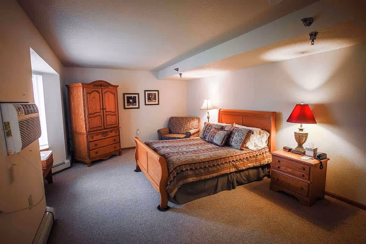 A cozy bedroom in a senior living facility featuring a wooden bed with patterned bedding and multiple pillows, a wooden nightstand with a red lampshade, a wooden armoire, a small armchair, two framed pictures on the wall, and a window air conditioning unit.