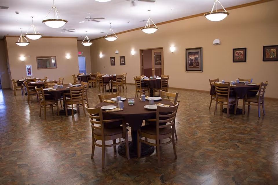 A spacious dining room with multiple round tables covered with dark tablecloths, each set with plates, cups, and utensils. Wooden chairs surround the tables. The room has beige walls adorned with framed artwork and is illuminated by ceiling lights and wall sconces. The floor has a patterned design.