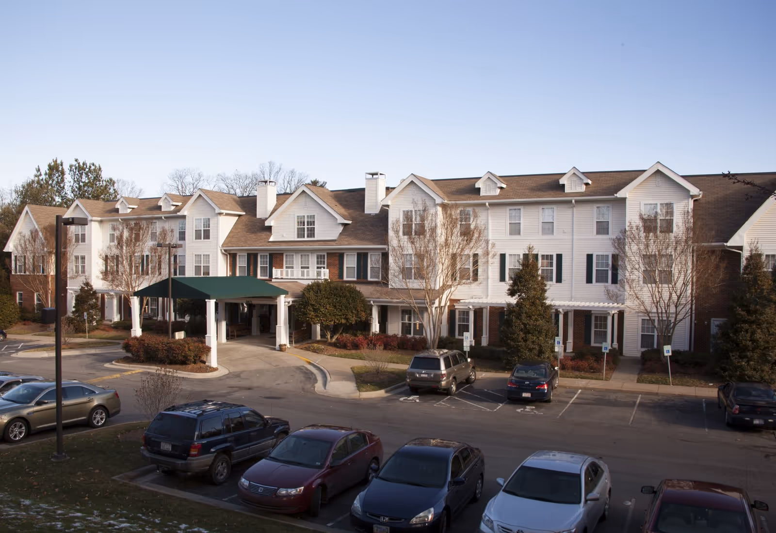 Front exterior of a multi-story senior living building with a covered entrance canopy and a parking lot with cars.