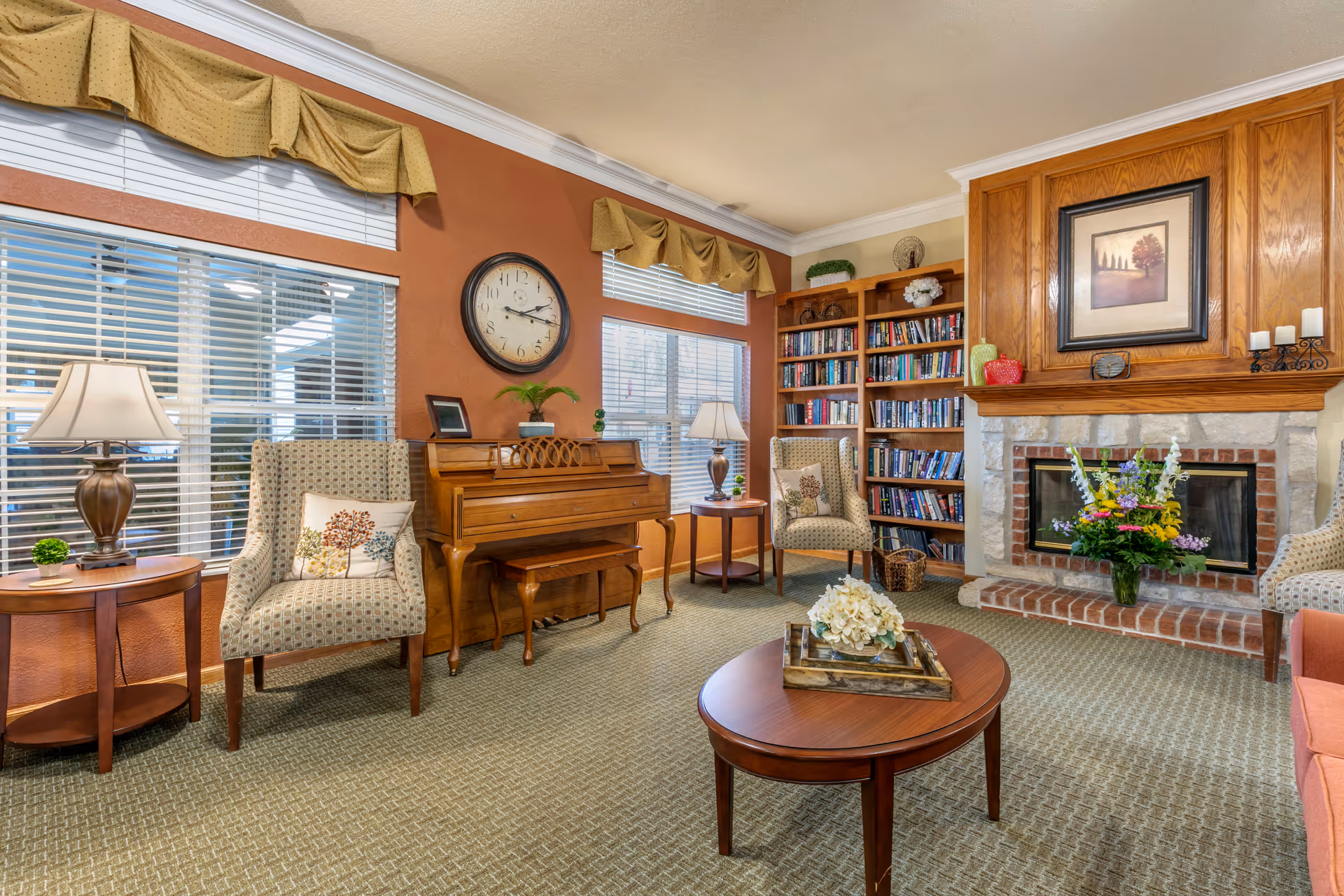 A cozy common living room with a wooden piano, armchairs, bookshelves, a fireplace with flowers, and a central coffee table.