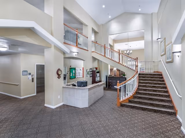 Interior view of a senior living facility lobby with a carpeted floor, a curved staircase with wooden handrails and white balusters, a reception desk with a computer, and beige walls with framed pictures and wall sconces.