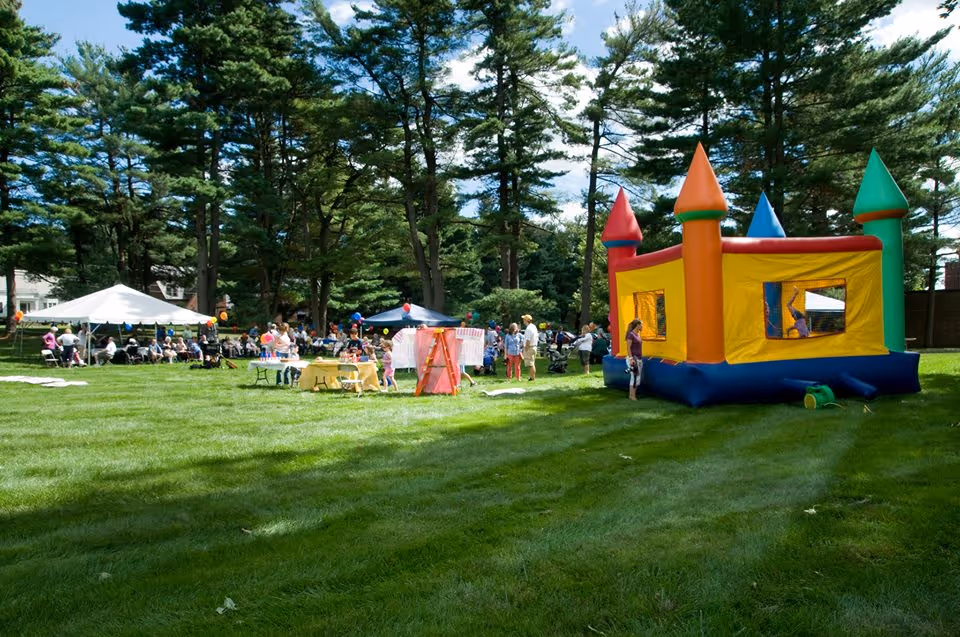 Outdoor scene at The Mercy Community with a large grassy area surrounded by tall trees. People are gathered under white and blue tents, with tables set up and children playing near a colorful inflatable bounce house shaped like a castle.