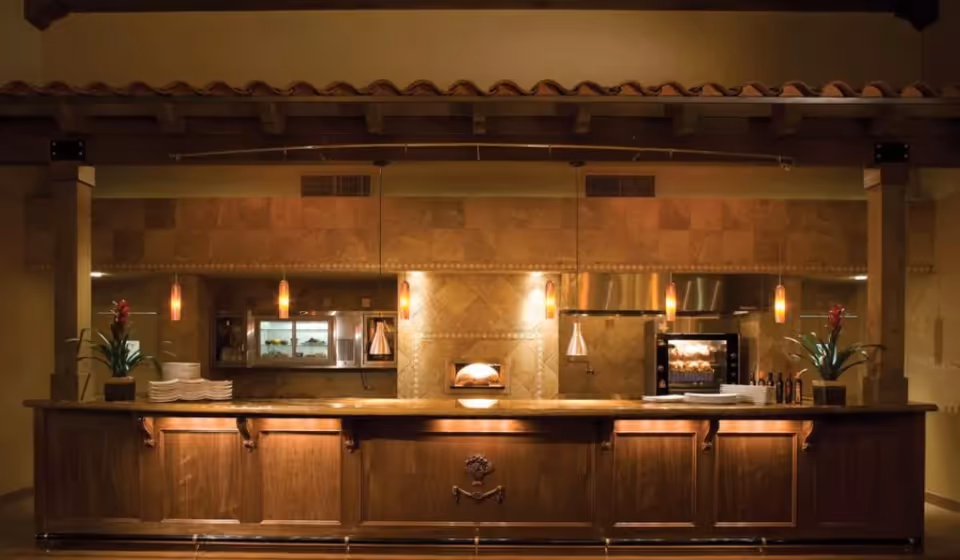 A warm, inviting kitchen area with a long wooden counter, pendant lights hanging from the ceiling, and kitchen appliances including an oven and a rotisserie. Two potted plants and stacks of plates are placed on the counter.