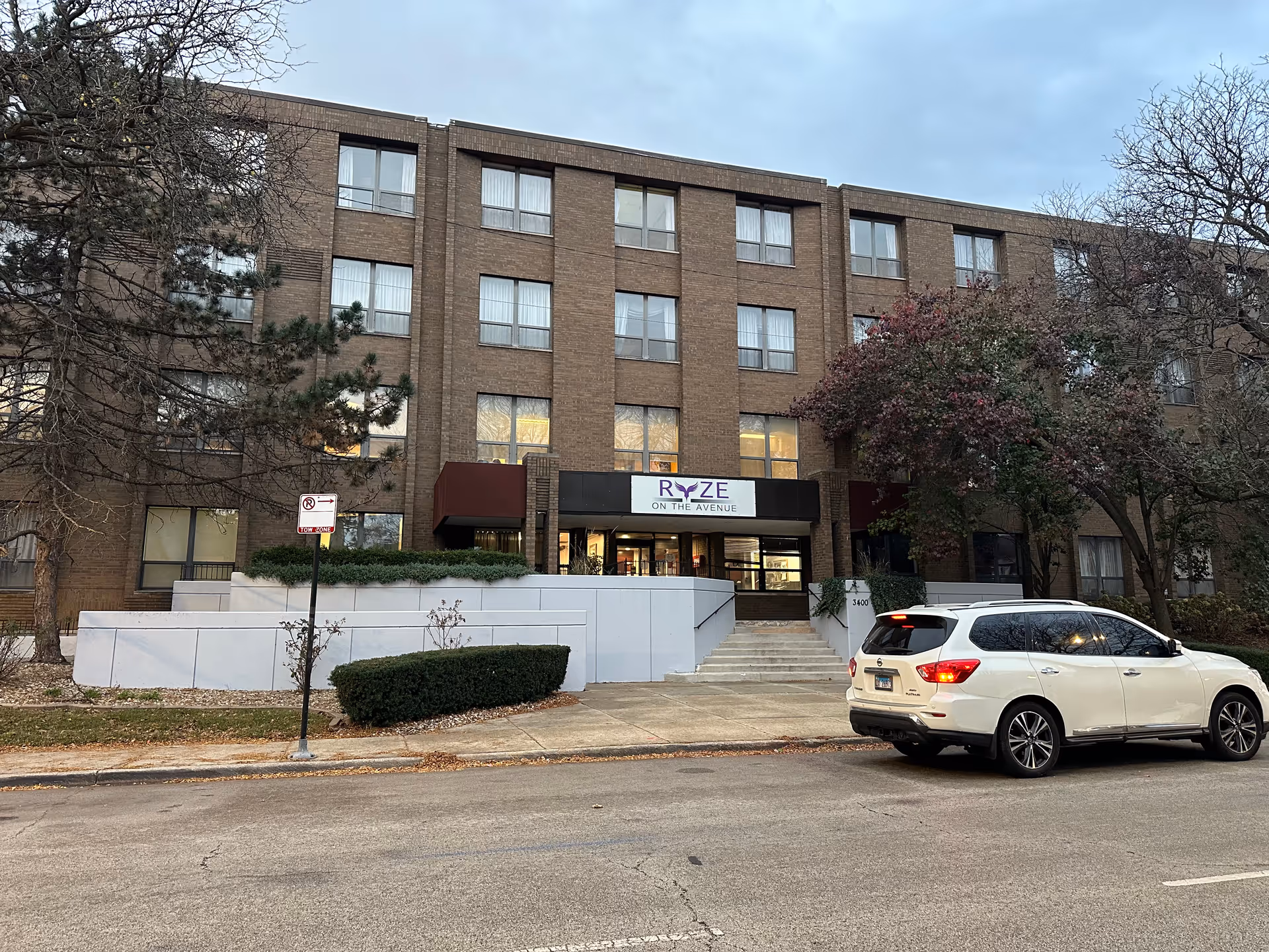 Exterior view of a four-story brick building with multiple windows and a main entrance with a sign above it that reads 'RYZE ON THE AVENUE'. There is a white SUV parked on the street in front of the building, and trees with sparse leaves surround the area under a cloudy sky.