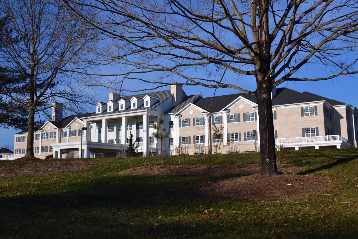 Exterior view of a large, multi-story senior living facility building with beige brick walls, white columns, and multiple windows. The building is set against a clear blue sky with leafless trees in the foreground and a grassy area with mulch around the trees.