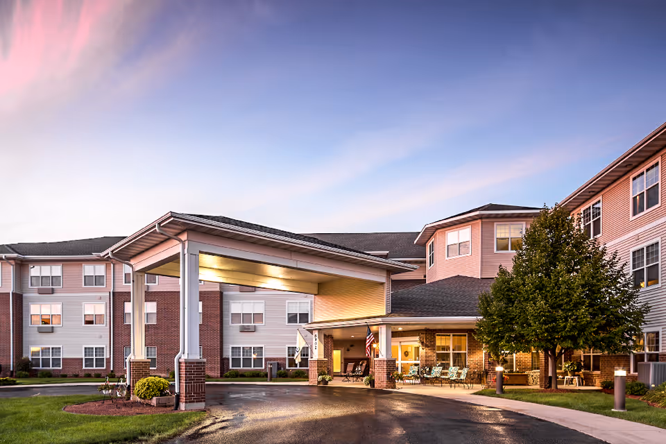 Exterior view of a senior living facility at dusk with a covered entrance, well-lit porch area, and a tree on the right side. The building has multiple windows and a combination of brick and siding on the facade.