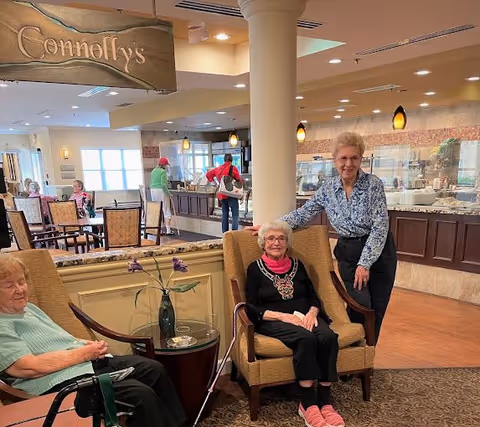 A dining area in a senior living facility with three elderly women, two seated in armchairs and one standing beside them. In the background, there is a buffet counter with food and people serving themselves. The area is well-lit with ceiling lights and pendant lamps. A sign above reads 'Connolly's'.