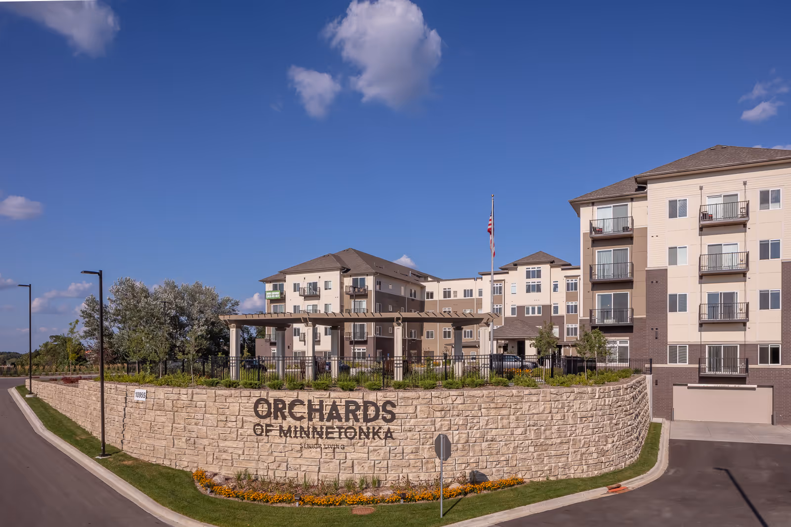 Exterior view of Orchards of Minnetonka Senior Living facility showing a multi-story building with balconies, a stone retaining wall with the facility name, landscaped greenery, and a clear blue sky with a few clouds.