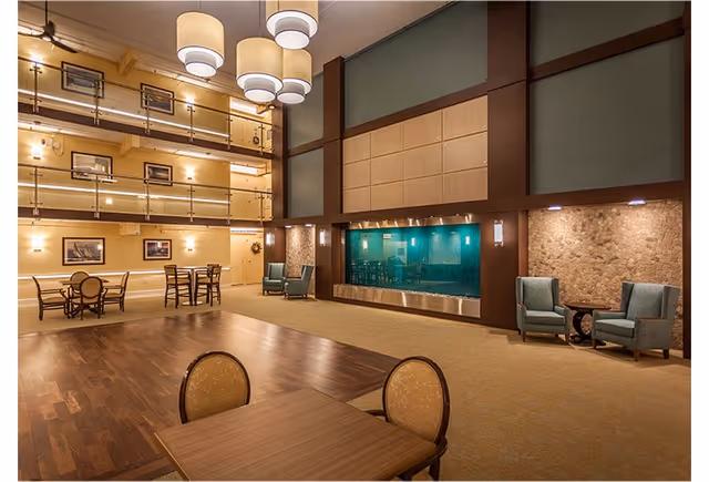 Spacious and well-lit interior common area with multiple seating arrangements including tables with chairs and armchairs. The room features a high ceiling with modern hanging light fixtures, a large decorative water feature on one wall, and framed artwork on the walls. The flooring is a combination of wood and carpet, and there is a balcony railing visible on the upper level.