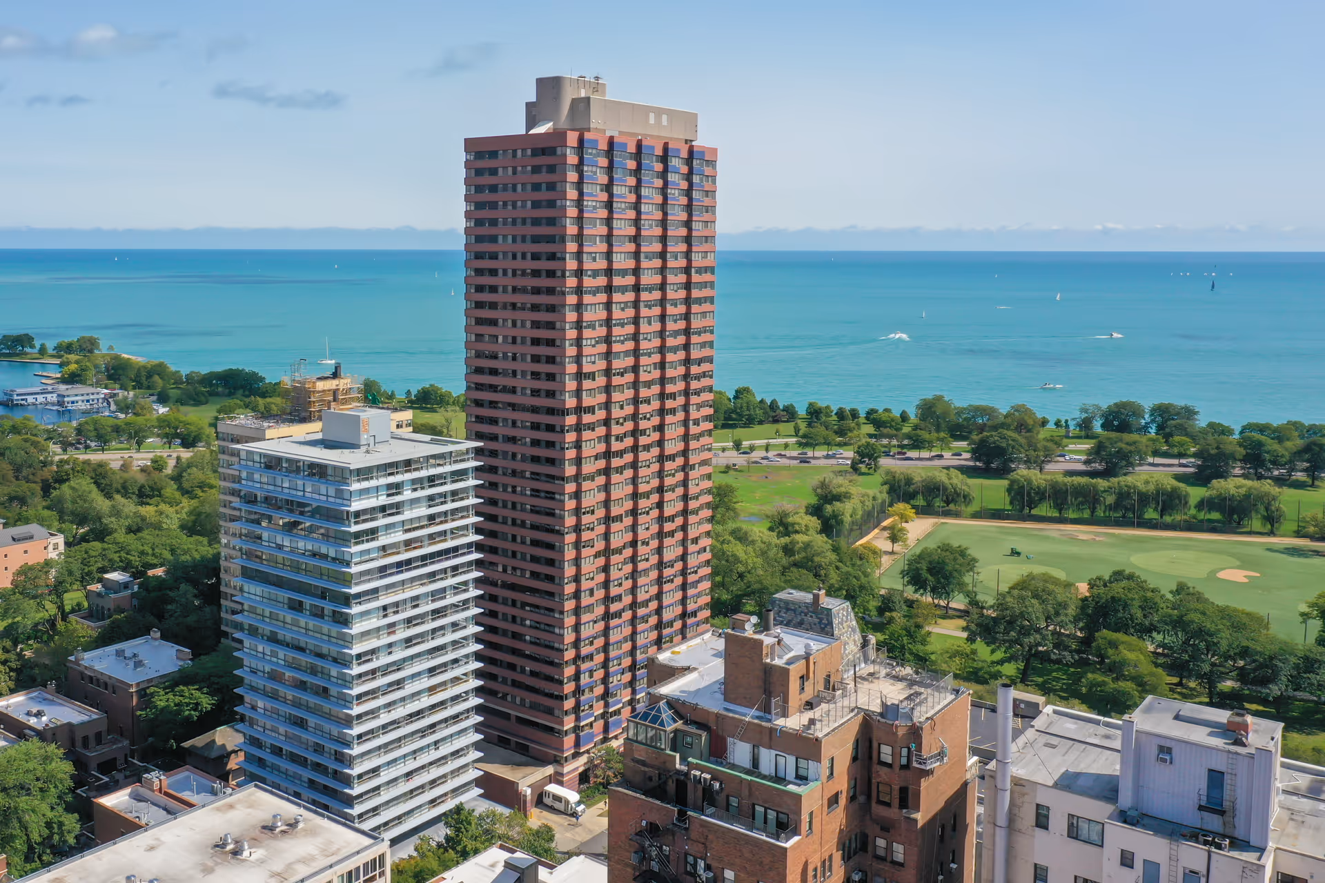 Aerial view of a tall residential building and surrounding smaller buildings near a large body of water with a park and golf course visible in the background under a partly cloudy sky.