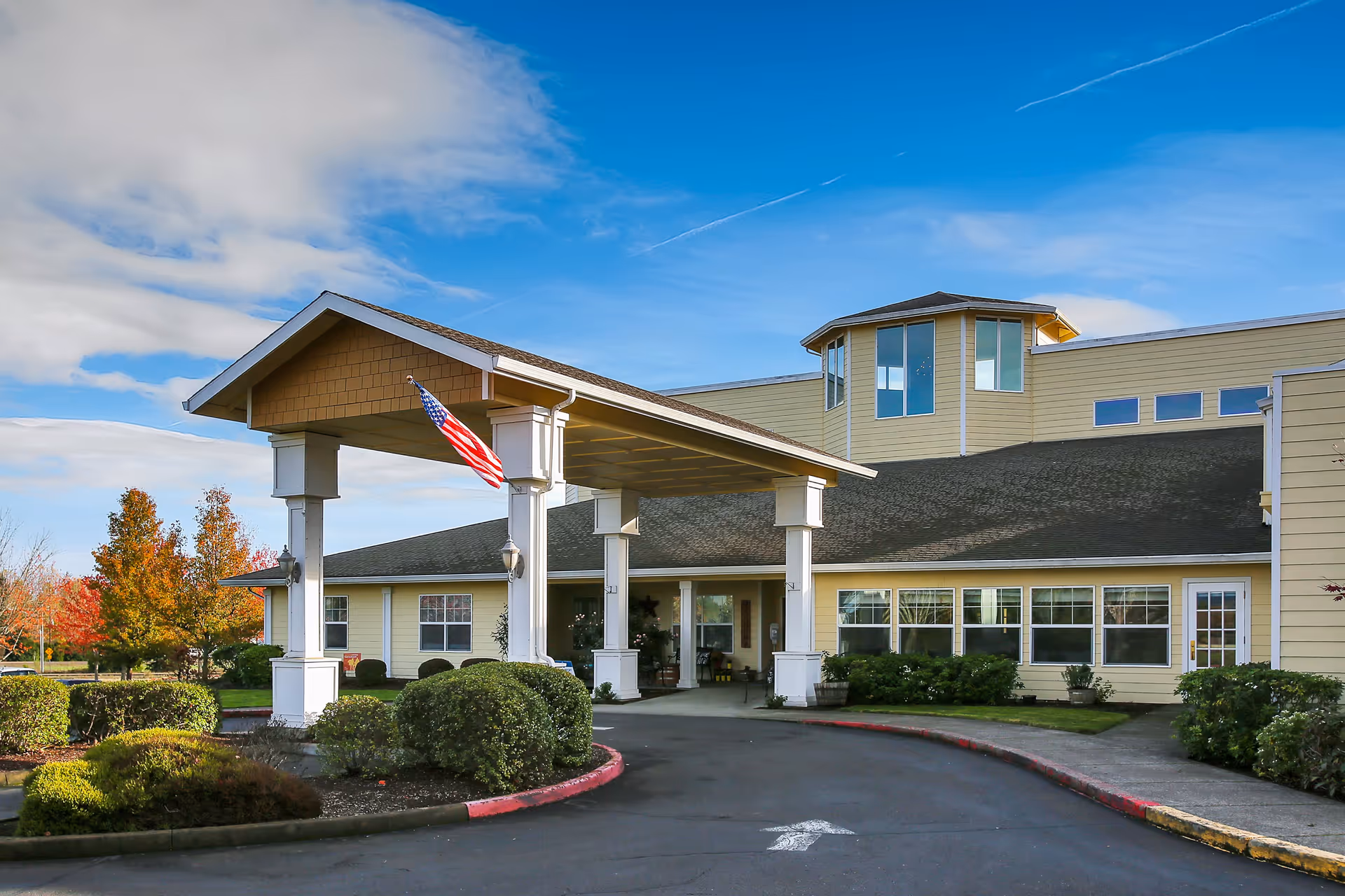 Exterior view of Prestige Senior Living West Hills building with a covered entrance supported by white columns, an American flag hanging, and landscaped bushes and trees under a partly cloudy blue sky.