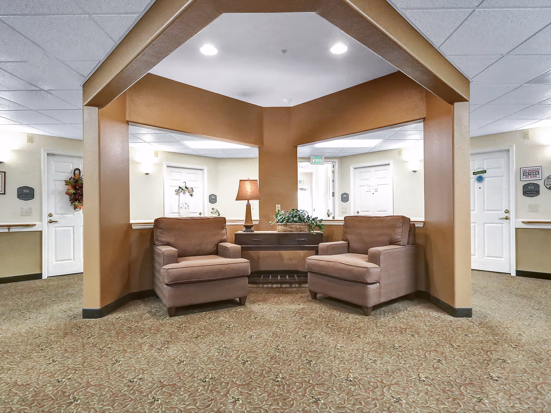 A seating area in a senior living facility hallway with two brown armchairs facing each other, a wooden side table with a lamp and a plant in between. The walls are beige with white doors labeled with room numbers, and the floor is covered with patterned carpet.