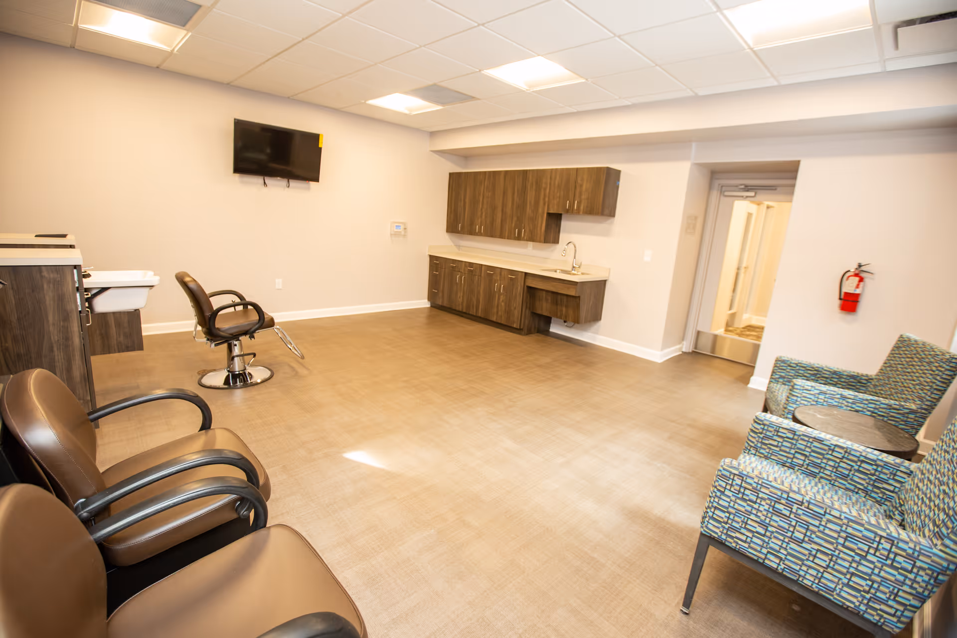 A spacious room in Norman Towers Senior Apartments featuring salon chairs, a wall-mounted TV, a sink with cabinetry, and a seating area with two patterned armchairs and a small round table. The room has a beige floor and light-colored walls with a ceiling of recessed lighting panels.