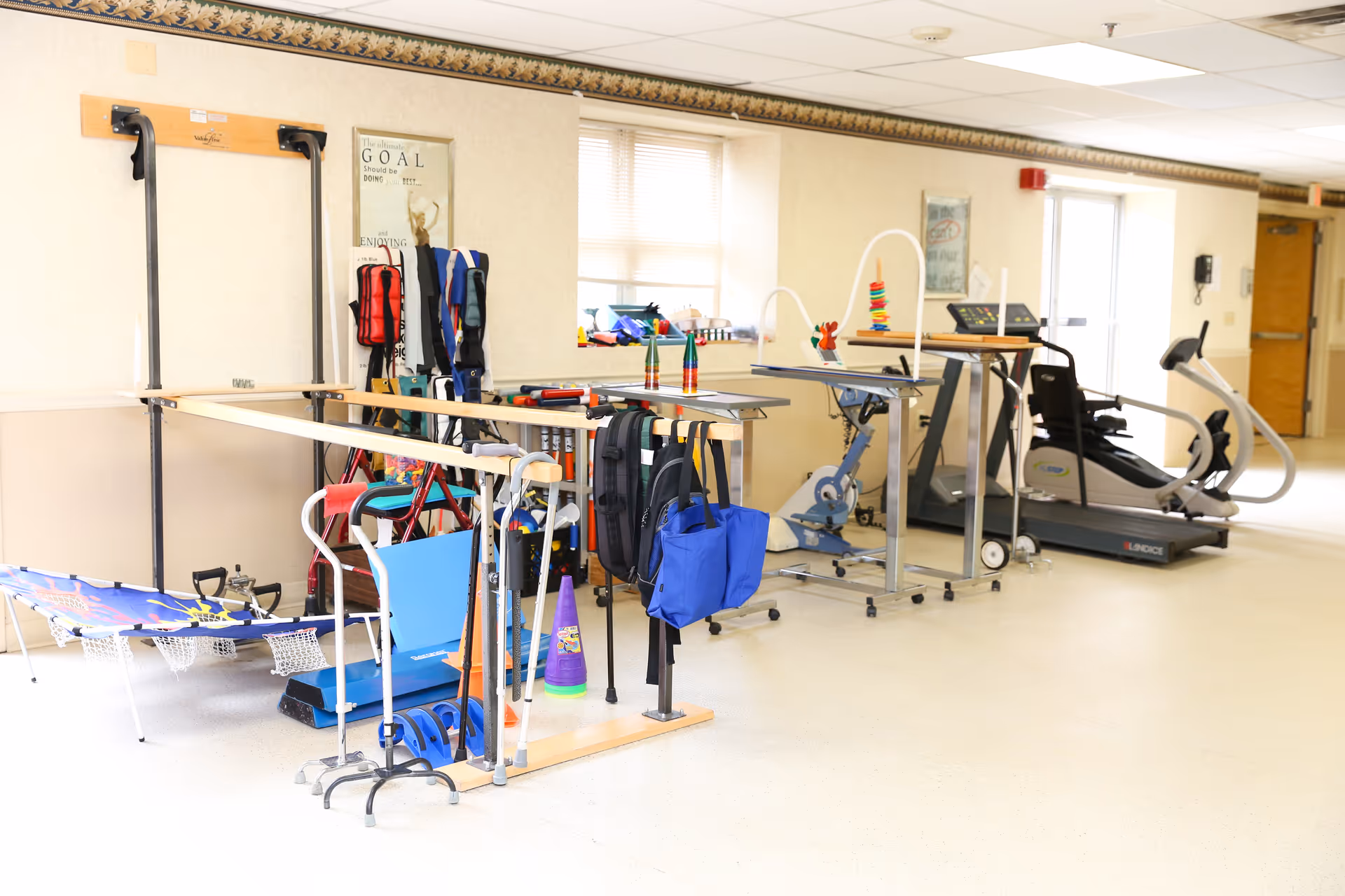 A bright rehabilitation or physical therapy room with various exercise and mobility equipment including parallel bars, walkers, a treadmill, a stationary bike, and therapy tools on tables. The room has large windows letting in natural light and motivational posters on the walls.