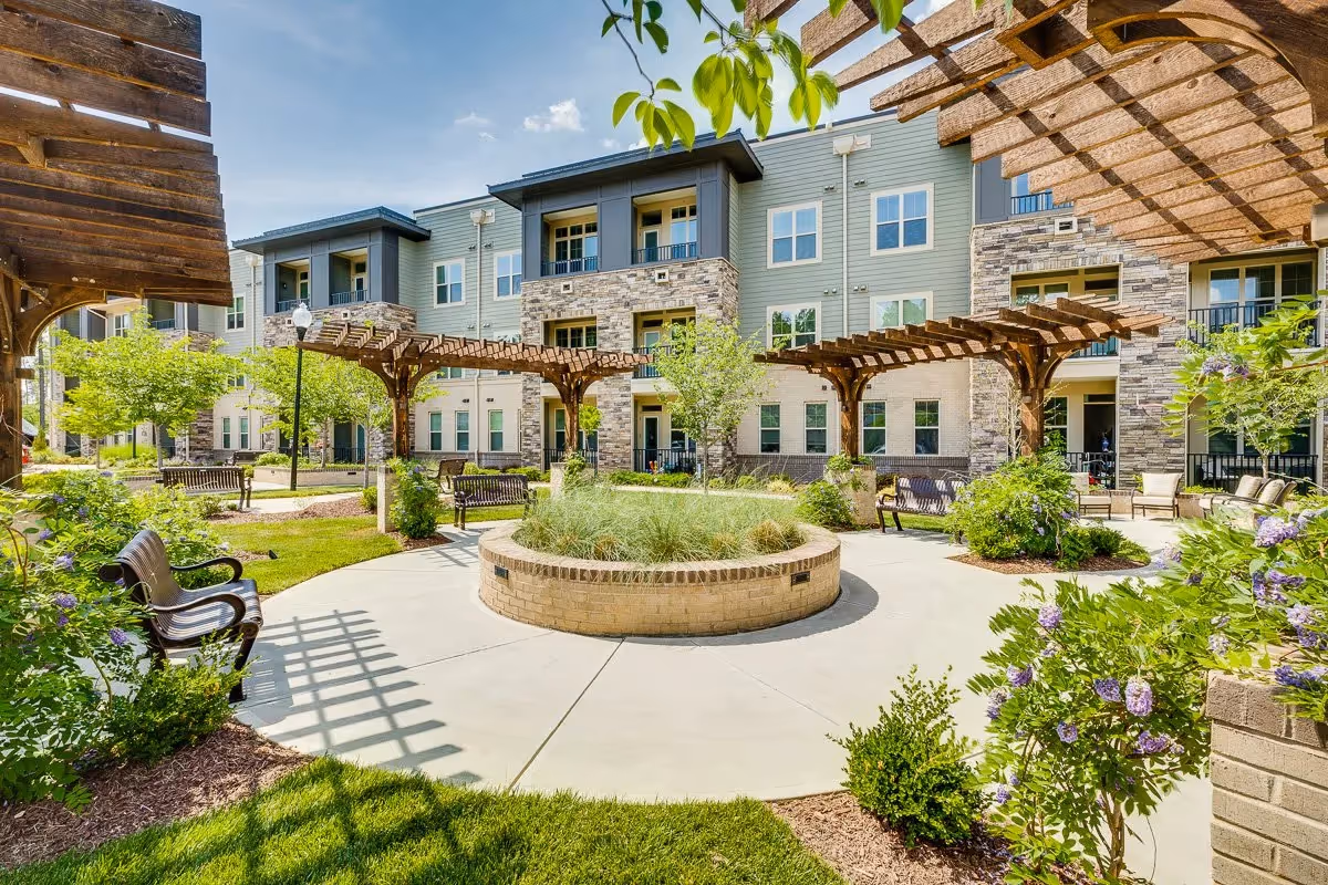 Outdoor courtyard area at The Reserve at Mills Farm featuring a circular paved walkway with a raised brick planter in the center, wooden pergolas providing partial shade, benches along the walkway, green grass, flowering bushes, and a multi-story residential building in the background under a blue sky.