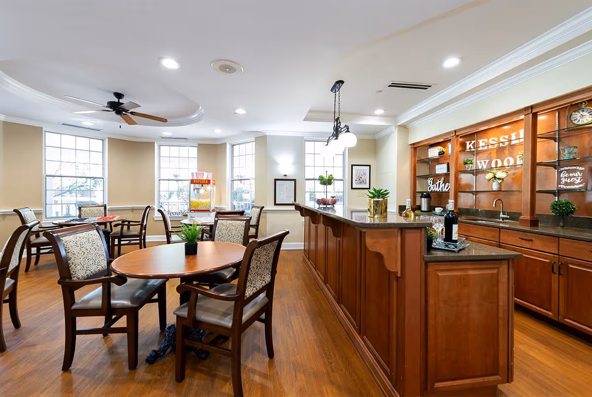 A bright and spacious common area with wooden floors and beige walls featuring multiple round tables with cushioned chairs. A wooden counter with a granite top is on the right side, decorated with plants, bottles, and a sink. Large windows allow natural light to fill the room, and a ceiling fan is mounted on the white ceiling. A popcorn machine is visible near the windows.