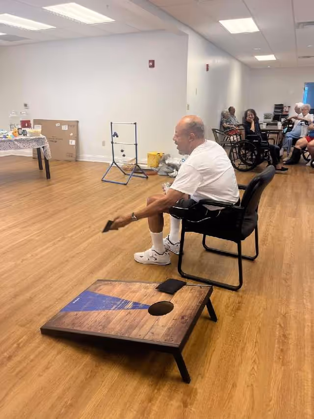 An elderly man sitting on a chair indoors playing a game of cornhole, aiming a bean bag at a wooden board with a hole. In the background, several other elderly people are seated, some in wheelchairs, watching and socializing in a spacious room with wooden flooring and white walls.