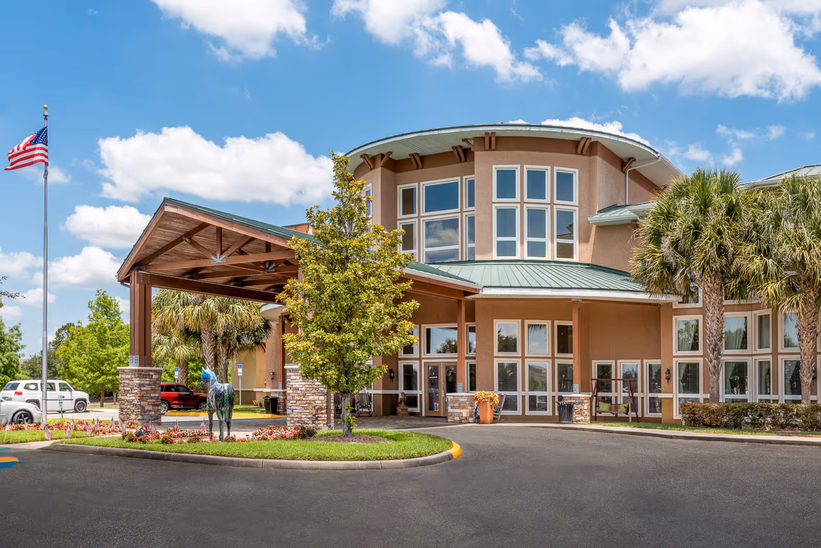 Exterior view of Brentwood at Fore Ranch senior living facility showing the main entrance with a covered drop-off area, surrounded by trees and landscaping under a blue sky with scattered clouds. An American flag is visible on a flagpole to the left.