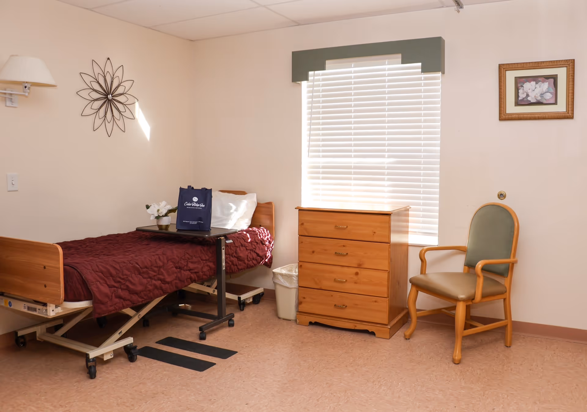 A simple senior living bedroom with a hospital-style bed covered with a maroon quilt, a small overbed table with a Cedar Ridge bag and a small flower pot, a wooden chest of drawers under a window with closed blinds, and a wooden chair with green upholstery. The walls are light-colored with minimal decoration including a metal wall art and a framed picture.