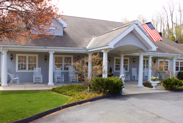 Exterior view of a single-story senior living facility building with a covered porch featuring white columns and several white rocking chairs. An American flag is mounted near the entrance. There are shrubs and a small tree in front of the building, with a paved driveway and a well-maintained lawn.