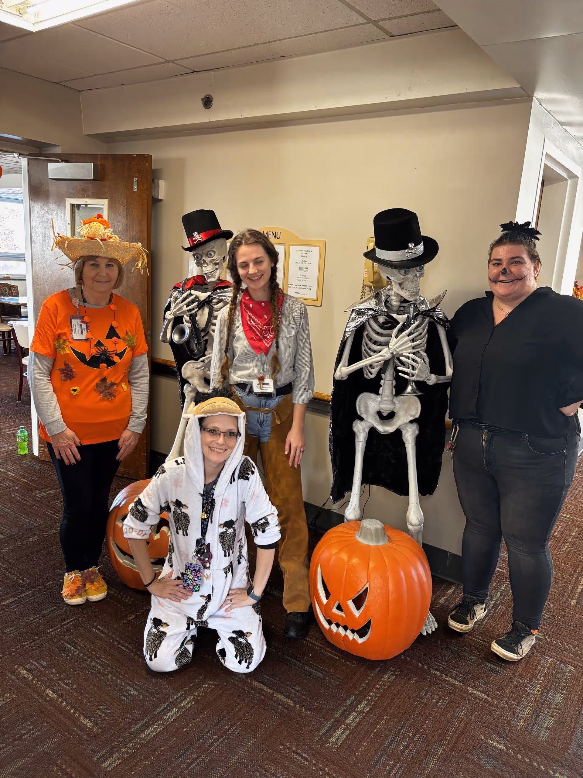 Five people dressed in Halloween costumes posing indoors next to two skeleton decorations and a large plastic jack-o'-lantern pumpkin. The costumes include a scarecrow, a cowgirl, a sheep onesie, and a black outfit with face paint. The setting appears to be a senior living facility hallway with a menu board on the wall.