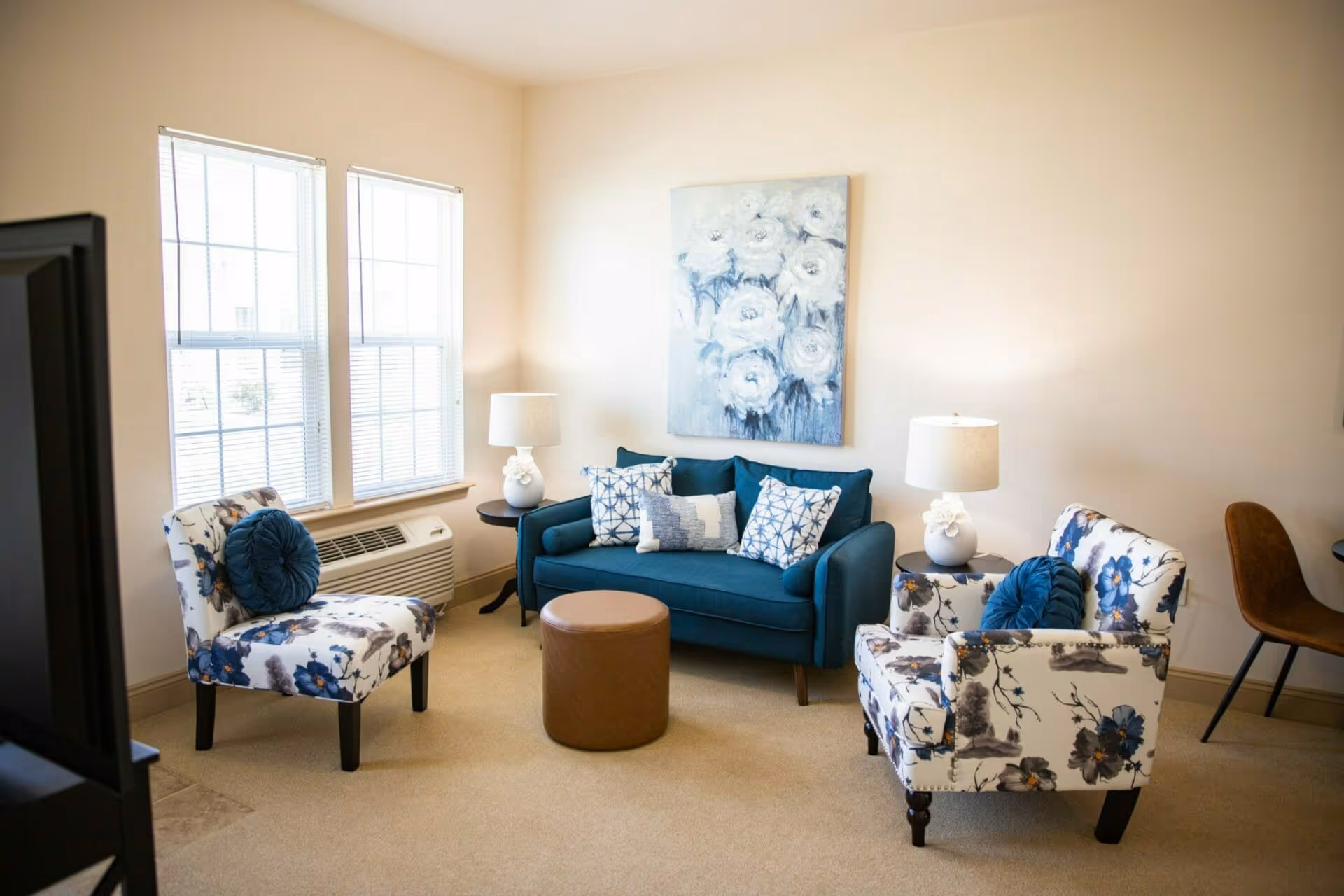 Bright seating area with a teal sofa, two floral armchairs, a round ottoman, lamps, and floral wall art.