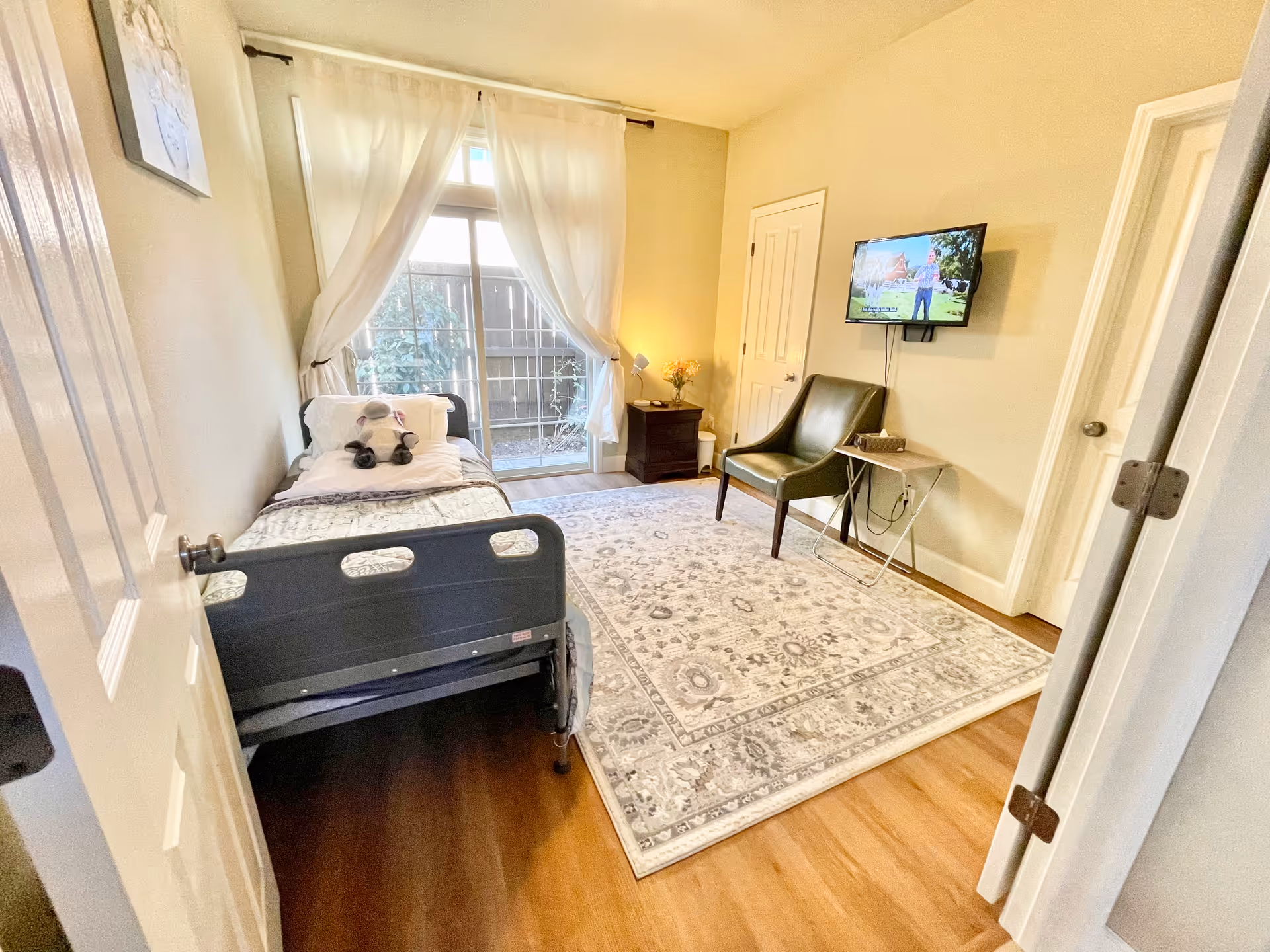 A bright and cozy bedroom with a hospital-style bed featuring a stuffed animal on the pillow. The room has light-colored walls, a large window with sheer white curtains letting in natural light, a patterned area rug on wooden flooring, a green chair, a small side table with a basket, a nightstand with a lamp and flowers, and a wall-mounted TV.