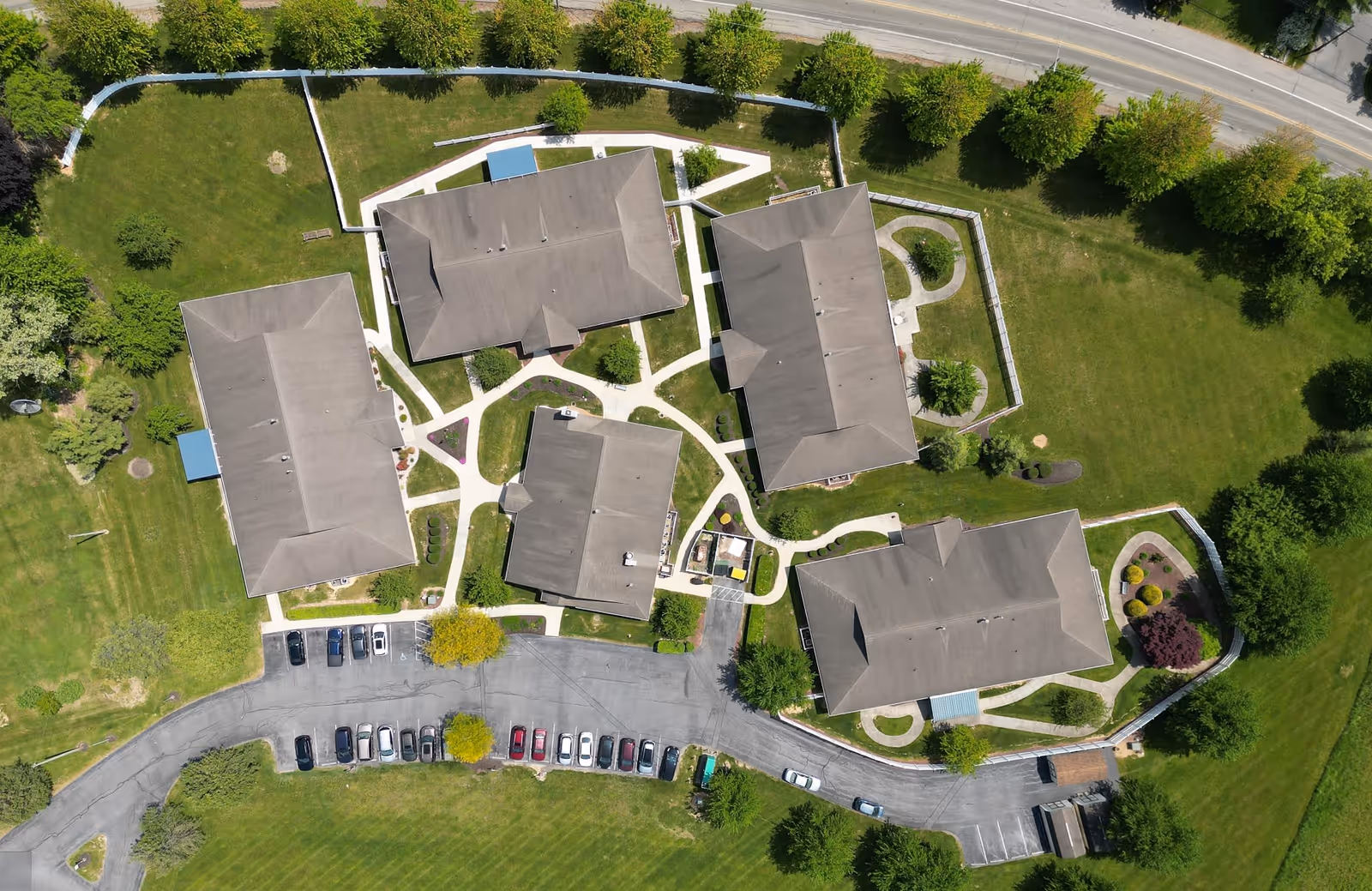 Aerial view of a senior living community with multiple buildings surrounded by green lawns, trees, and walking paths. There is a parking area with several cars parked along a curved driveway.