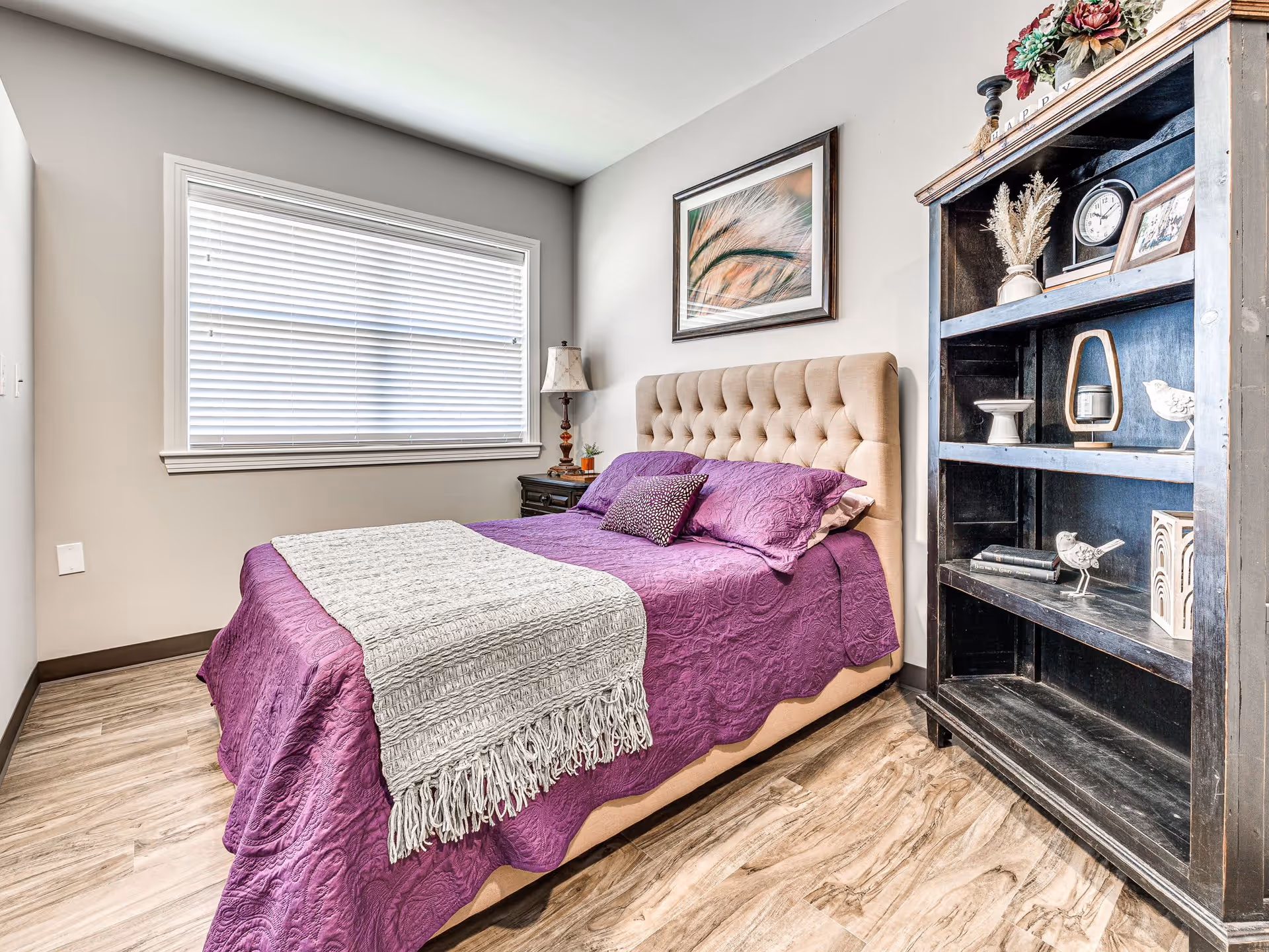 Well-lit bedroom with a tufted headboard bed dressed in purple bedding, a nightstand and a dark wooden bookshelf against a neutral wall.