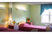 A senior living facility bedroom with two single beds covered in purple bedspreads. There are two wooden nightstands between and beside the beds, each with a lamp and a flower arrangement. The walls are painted light green, and there is a large window with blue curtains allowing natural light into the room.
