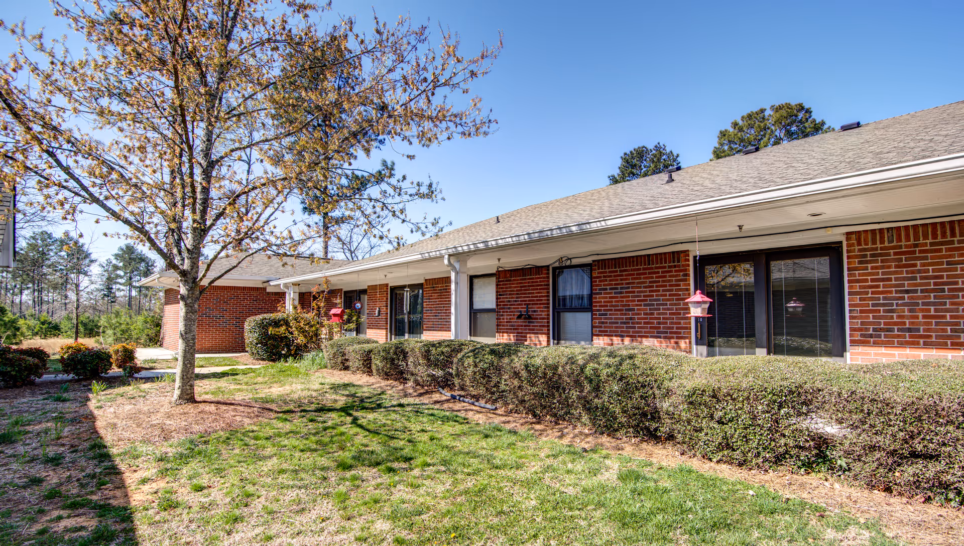 Exterior view of a single-story brick building with a sloped roof, surrounded by green grass, bushes, and a tree with sparse leaves under a clear blue sky.