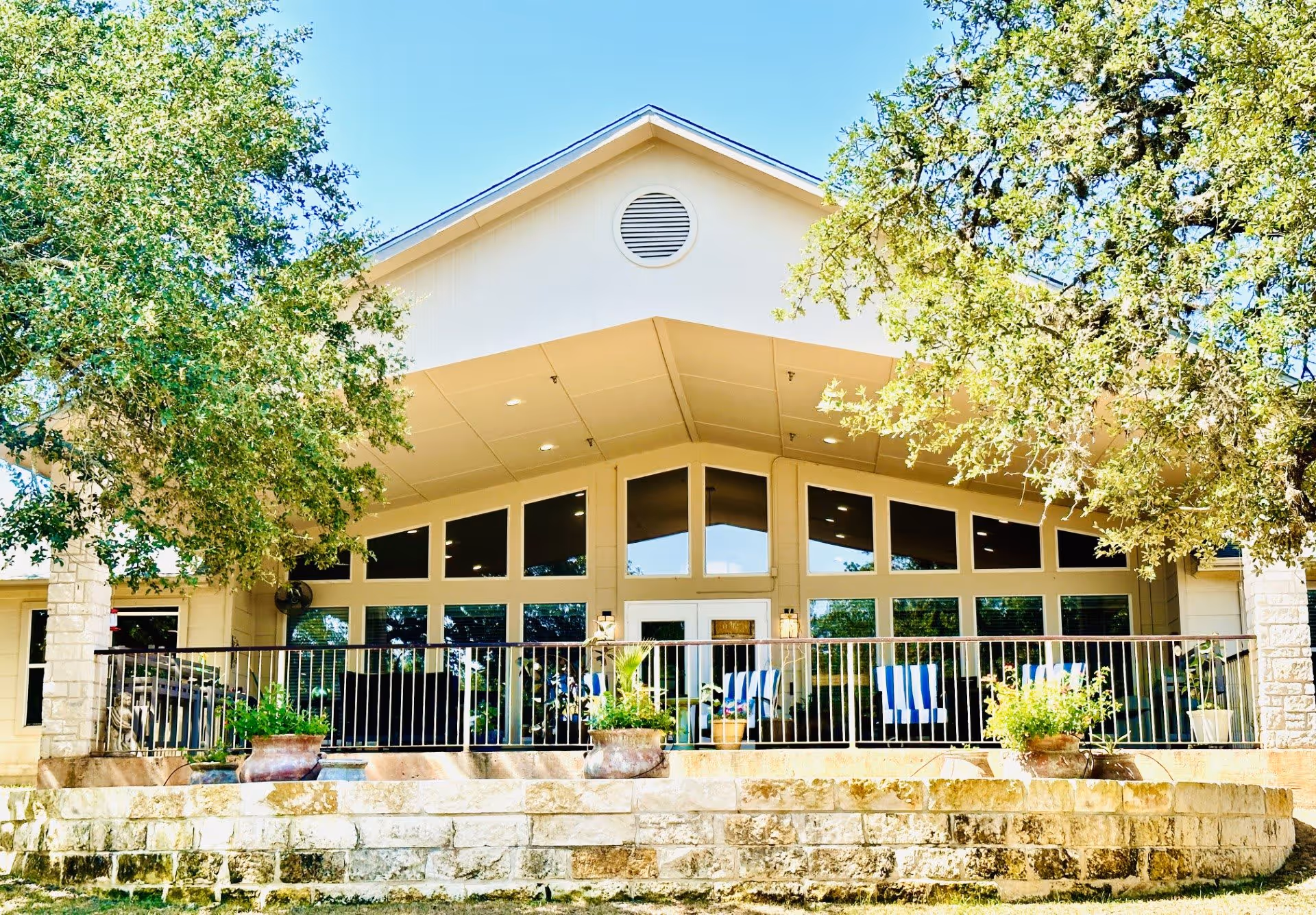 Exterior view of a building with large windows and a covered porch area featuring several chairs with blue and white striped cushions. The building is surrounded by trees and has a stone retaining wall with potted plants in front.