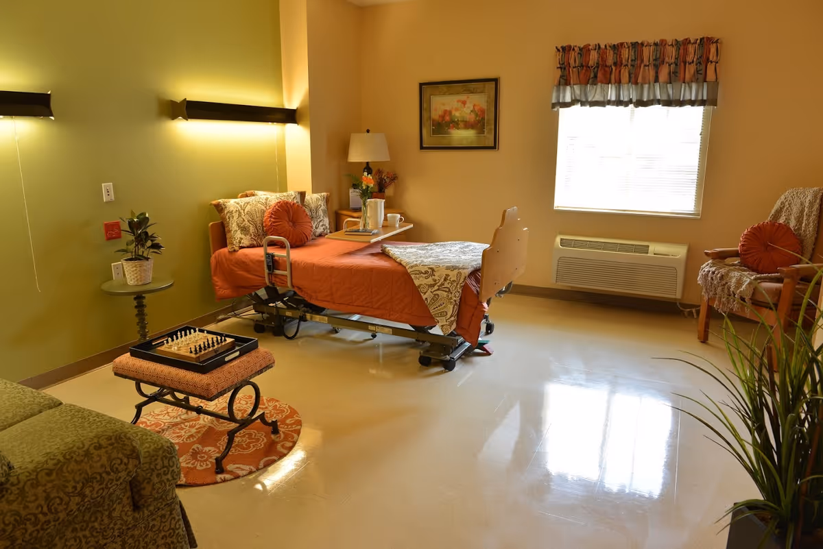 A cozy senior living bedroom with a hospital-style bed covered in orange and patterned bedding. The room has a green wall with two wall-mounted lights, a small round table with a potted plant, a chessboard on a cushioned stool, a window with a valance, an armchair with a throw blanket and orange pillow, a framed floral painting, and a floor plant near the foreground.