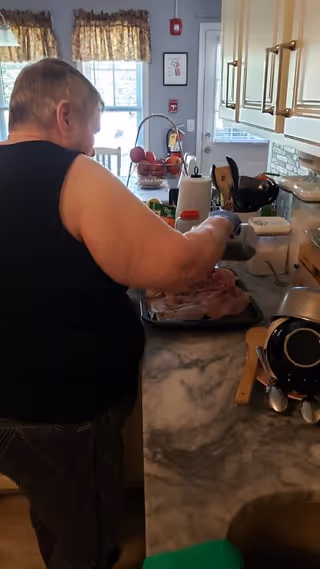 An elderly person wearing a black sleeveless top is preparing food on a kitchen counter. The kitchen has light-colored cabinets, a marble countertop, and various kitchen utensils and appliances. There is a window with floral curtains and a door in the background.