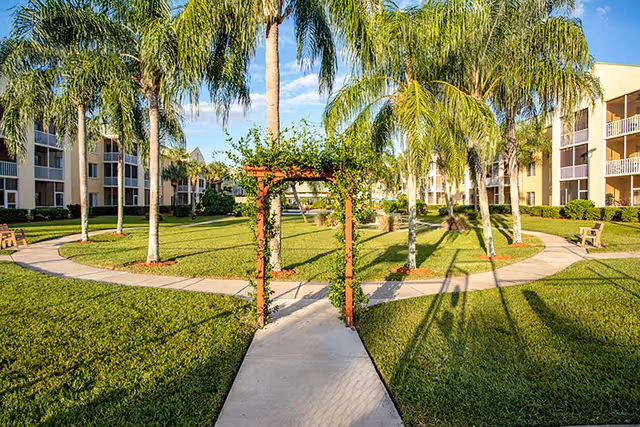 A sunny outdoor courtyard area at The Fountains of Melbourne featuring a concrete pathway leading through a wooden archway with climbing plants. The courtyard is surrounded by palm trees, green grass, benches, and multi-story residential buildings with balconies.