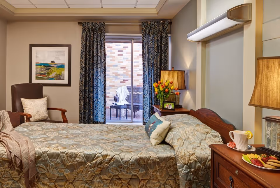 A cozy bedroom in a senior living facility with a neatly made bed covered in a patterned bedspread. There is a wooden nightstand with a lamp, a cup with a lemon slice, and a plate of fruit and cookies. A vase with orange tulips and a framed photo are also on the nightstand. A comfortable armchair with a pillow is positioned near a window with patterned curtains, which opens to a small outdoor patio with wicker chairs and a table.