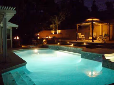 Nighttime outdoor pool area with lit water, torches, chairs, and a gazebo.